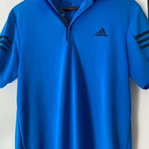 Blå pikétröja från Adidas - Snygg blå pikétröja från Adidas i tränings material, med korta ärmar och klassisk krage. Tröjan har Adidas-loggan på bröstet och tre ränder på ärmarna. Perfekt för en sportig look! Tveka inte på att höra av er vid frågor eller funderingar! Mvh Sixten 