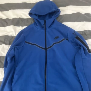 Blå hoodie från Nike - Säljer en blå hoodie från Nike med dragkedja och huva. Den har svarta detaljer och en ficka på ärmen. Perfekt för en sportig look eller avslappnade dagar.