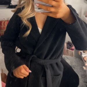 Svart kappa med bälte - Snygg svart kappa med bälte som ger en stilren look. Perfekt för höst och vår. Den har långa ärmar och en klassisk design som passar till många olika outfits.