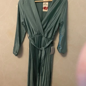 Hell ny Lång grön omlottklänning Wolfen - Säljer hell ny Snygg långklänning i grön satin från Wolfen. Klänningen har omlottmodell med v-ringning, långa ärmar och ett knytband i midjan. Kjolen är plisserad för extra volym och flow. Perfekt för dig som vill sticka ut med stilren elegans. Storlek S/M