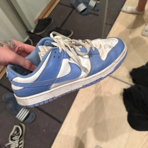 Nike Dunk Low blå och vita sneakers - Nike Dunk Low sneakers i blått och vitt skinn med klassisk swoosh på sidorna. Lågt skaft, vita snören och perforerad tåbox för extra ventilation. Perfekt för dig som gillar retrostil och streetwear. passar dig som har storlek 43 med breda fötter också