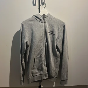 Grå hoodie från Peak Performance - Säljer en stilren grå hoodie från Peak Performance med dragkedja framtill och två fickor. Tröjan har huva och diskret logga på bröstet. Perfekt för chill dagar eller när du vill ha en enkel look.
