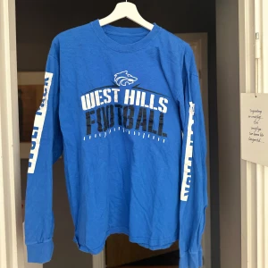 Blå långärmad West Hills Football-tröja - Blå långärmad tröja med tryck 'West Hills Football' framtill och text på ärmarna. Crewneck-modell i bomull, perfekt för dig som gillar sportig och avslappnad stil. Snygg att matcha med jeans eller joggers.