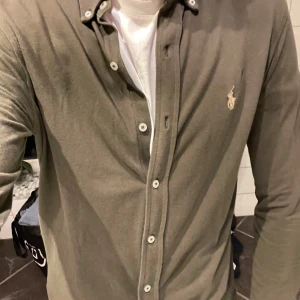 Olivgrön skjorta från Ralph Lauren - Snygg olivgrön skjorta från Ralph Lauren i lätt och luftig bomullskvalitet. Klassisk button-down-krage, vita knappar och ikonisk broderad logga på bröstet. Perfekt för dig som gillar stilrena och tidlösa plagg.