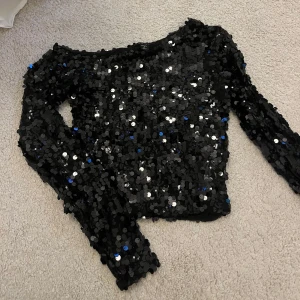 Svart paljettopp från newyorker - Glittrig svart topp från newyorker, täckt med stora paljetter i svart. Modellen är croppad med långa ärmar och rund halsringning. Perfekt för dig som vill sticka ut och glänsa extra mycket. Använd endast 2 gånger.