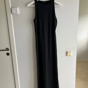 Svart långklänning från Gina tricot - En stilren svart långklänning med ärmlös design och rund halsringning. Klänningen har en rak och enkel passform som ger en clean look, perfekt att styla med sneakers eller boots. Materialet känns mjukt och följsamt mot kroppen.