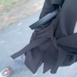 Svart kappa med bälte  - Svart lång kappa från Amisu i klassisk modell med bred krage och knytskärp i midjan. Kappan har slits bak och raka, långa ärmar. Perfekt för dig som vill ha en stilren och tidlös look under höst och vinter.