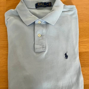 Ljusblå pikétröja från Ralph Lauren - Säljer en klassisk ljusblå pikétröja från Ralph Lauren med broderad logga på bröstet. Tröjan har krage, två knappar och korta ärmar. Perfekt för en clean och stilren look. Materialet är mjuk bomull som känns skönt mot huden.