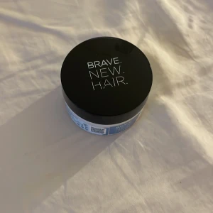BRAVE.NEW.HAIR hårmask - En rund burk med svart lock och vit text från BRAVE.NEW.HAIR. Innehållet är en vit, krämig hårmask. Perfekt för att ge håret extra fukt och näring. Snygg och stilren design som passar på badrumshyllan. Aldrig använd!