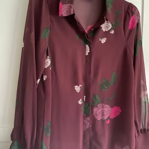 Blommig vinröd skjorta från OBJECT - Vinröd skjorta från OBJECT med blommigt mönster i rosa, lila och grönt. Skjortan har klassisk krage, knappar framtill och långa ärmar med lätt puff vid ärmslut. Tunt och flowy material som ger en snygg siluett.