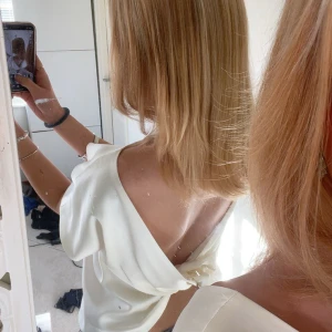 Vit omlottopp i satin med öppen rygg - Säljer en vit omlottopp i satin med korta ärmar och snygg öppen rygg. Toppen har en elegant knutdetalj bak som ger en unik look. Perfekt för dig som gillar stilrena och trendiga plagg med det lilla extra.