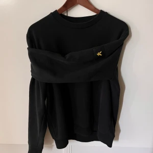 Svart sweatshirt från Lyle & Scott - Svart sweatshirt från Lyle & Scott i relaxed fit. Tröjan har rund halsringning, långa ärmar och klassisk gul logga på bröstet. Perfekt basic-plagg med stilren look och mjukt material som passar till allt.
