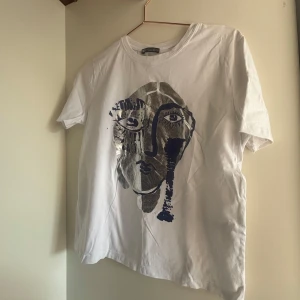 Vit t-shirt med konstnärligt tryck Zara - Vit t-shirt från Zara med ett stort konstnärligt ansikte i silver och mörkblått på framsidan. Klassisk rund hals och korta ärmar. Skön och mjuk bomullskvalitet, perfekt för en cool och avslappnad stil.