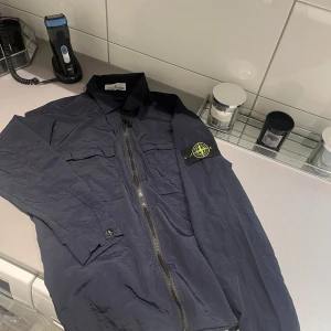 Stone Island overshirt i mörkblått - Säljer en mörkblå overshirt från Stone Island med klassisk patch på ärmen. Jackan har två bröstfickor med lock, hel dragkedja framtill och lång ärm. Väldigt tunn så perfekt för sommarskvällar. 