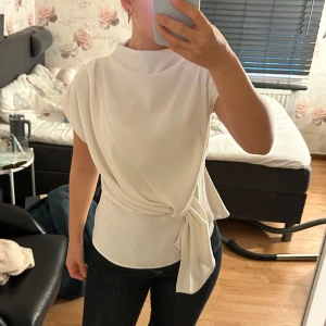 Vit omlottopp med knyt i midjan - Säljer en stilren vit omlottopp med hög hals och knyt i midjan. Toppen är ärmlös och har en snygg drapering framtill som ger en cool och modern look. Perfekt att matcha med jeans eller kjol för en clean outfit.
