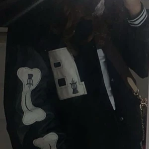 Svart bomberjacka med benpatches - Säljer en svart oversize bomberjacka med vita detaljer och stora benpatches på ärmarna. Jackan har en stor vit bokstavspatch på bröstet och randiga muddar vid ärmslut och krage. Perfekt för dig som vill sticka ut med en unik streetstyle.