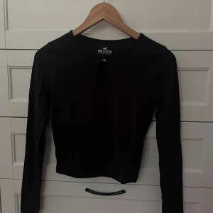 Svart långärmad topp från Hollister - Svart långärmad topp från Hollister Must-Have Collection. Toppen har en rund halsringning och en snygg droppformad cut-out framtill. Modellen är croppad och sitter tajt mot kroppen, perfekt att styla med höga jeans eller kjol.