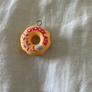 Donut-halsband med text och strössel. - Supergulligt halsbandshänge i form av en donut med gul bas, färgglada strössel och texten 'CHOCOLATE' i rosa. Hängsmycket har en rund form och en liten ögla för kedja. Perfekt för dig som gillar söta och lekfulla accessoarer.