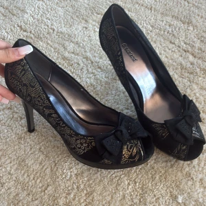 Svarta pumps med rosett från Graceland - Snygga svarta pumps från Graceland med hög klack och öppen tå. Skorna har ett elegant spetsmönster i guld och en stor svart rosett framtill med små glittriga detaljer. 