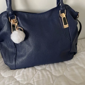 Mörkblå axelväska med gulddetaljer - Snygg mörkblå axelväska i syntet med guldfärgade detaljer och en fluffig vit nyckelring. Väskan har struktur i materialet, dragkedja på baksidan och avtagbar axelrem. Perfekt storlek för att få plats med allt du behöver.