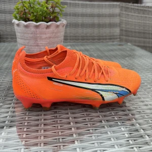 Orangea Puma fotbollsskor med snörning - Snygga orangea fotbollsskor från Puma med svart och vit logga på sidan och blå detaljer. Skorna har snörning, platt sula med fasta dobbar och är tillverkade i syntetmaterial med stickad ovandel för extra komfort och passform. Perfekta för dig som vill sticka ut på planen.