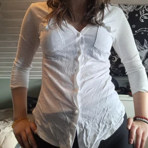 Vit v-ringad skjorta med knappar - Säljer en vit, v-ringad skjorta i tunn bomull med knappar framtill och två bröstfickor. Skjortan har trekvartsärm och en figurnära passform som framhäver siluetten. Perfekt att styla öppen eller stängd beroende på humör. Den har även ribbade sidor.