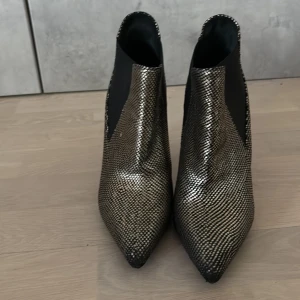 Metallicglittriga stövletter med spetsig tå - Snygga stövletter från Saint Laurent i metallicglittrigt mönster med svart resår på sidorna. Skorna har spetsig tå och hög smal klack, perfekt för dig som vill sticka ut. Materialet har en metallic-finish och reptil-inspirerat mönster som ger en edgy vibe. 