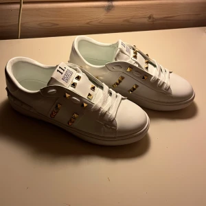Valentino Rockstud sneakers vit/guld - Snygga vita Valentino Rockstud Untitled sneakers med ikoniska guldfärgade nitar och klassisk låg siluett. Skorna har vita snören, platt sula och är tillverkade i skinn. Perfekta för dig som vill sticka ut med exklusiva detaljer och lyxig känsla.