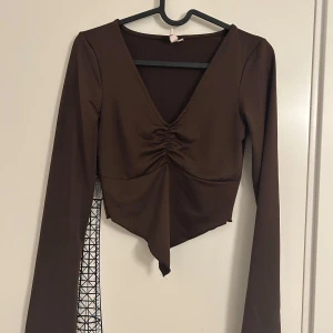 Brun långärmad topp med rynkdetalj från Nelly  - Brun långärmad topp med v-ringning och snygg rynkdetalj framtill från Nelly i storlek XS. Toppen har en asymmetrisk nederkant och en figurnära passform. 