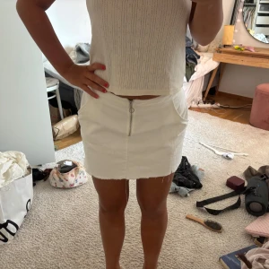 Vit jeanskjol med dragkedja - Säljer en snygg vit kort jeanskjol med rå kant och silverfärgad dragkedja framtill. Kjolen har fickor på sidorna och en clean, enkel look som är lätt att matcha med olika toppar. Perfekt för dig som gillar en fräsch och stilren vibe.