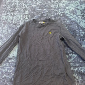 Svart långärmad tröja Lyle & Scott - Svart långärmad tröja från Lyle & Scott med rund hals och klassisk gul logga broderad på bröstet. Tillverkad i mjuk bomull som är skön mot huden. Perfekt för dig som gillar stilrena och enkla plagg med en sportig touch.