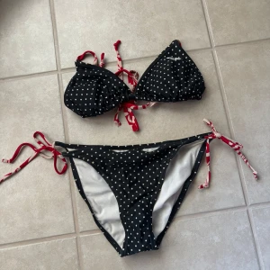 Prickig bikini från Roxy med knyt - Svart bikini från Roxy med vita prickar och röda knytband med mönster. Triangeltopp och trosa med sidor som knyts, båda i mjukt syntetmaterial. Perfekt för stranden och sommarens bad.