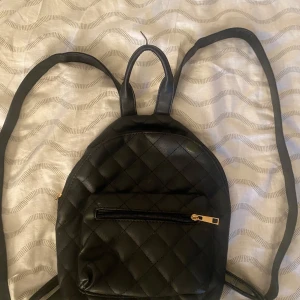 Svart quiltad ryggsäck i skinnimitation - Säljer en svart quiltad ryggsäck i skinnimitation med guldiga dragkedjor. Väskan har ett stort huvudfack och en mindre ficka framtill, perfekt för mobilen eller nycklar. Stilren och enkel design som passar till det mesta.