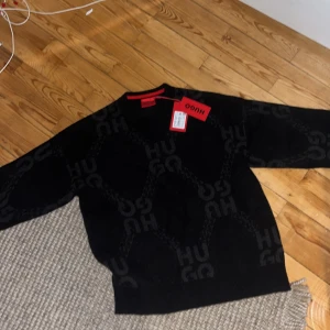 Svart stickad tröja från Hugo boss  - Säljer en svart stickad tröja från Hugo med diskret logomönster över hela tröjan. Tröjan har rund halsringning med röd detalj och långa ärmar. Perfekt för dig som gillar stilrena plagg med en twist. Storlek S lite stor i storleken, aldrig använt pris lapp även kvar.