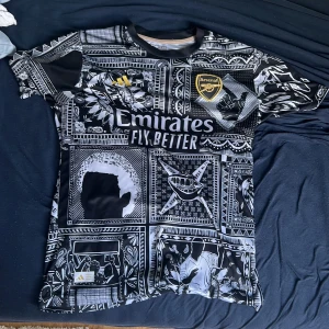 Arsenal Saka 87 svartvit fotbollströja - Unik Arsenal fotbollströja med Saka 87 på ryggen. Svartvit med grafiskt, rutigt mönster och detaljerade tryck. Adidas-logga och Emirates-sponsor på bröstet, Arsenal-märke i guld. Kortärmad och tillverkad i lätt, andasvänligt material.