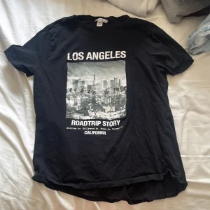 Svart t-shirt med LA-print från Amisu - Svart t-shirt från Amisu med tryck av Los Angeles och texten 'Roadtrip Story California' på framsidan. Klassisk rund hals och korta ärmar. Mjuk och skön i bomull, perfekt till jeans eller shorts för en avslappnad vibe.