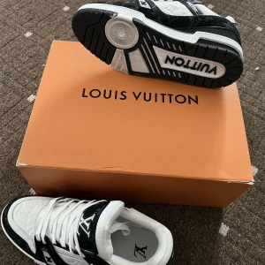 Louis Vuitton LV Trainer Sneaker svart/vit - Louis Vuitton LV Trainer Sneaker i svart och vitt med tydliga LV-loggor och monogramdetaljer. Skorna har snörning, platt sula och är tillverkade i en mix av skinn och textil. Snygg kontrast mellan svart och vitt, perfekt för dig som gillar exklusiva sneakers med statement.