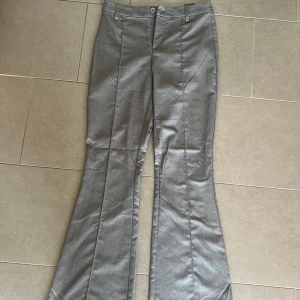 Grå bootcut kostymbyxor H&M Divided - Snygga grå kostymbyxor från H&M Divided i bootcut-modell. Byxorna har pressveck, bälteshällor och stängs med knapp och dragkedja. Perfekta för en clean och stilren look. Materialet är mjukt och har en lätt glansig finish.