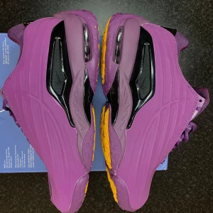 Lila Nike NOCTA sneakers - Unika Nike NOCTA sneakers i lila med svarta och gula detaljer. Skorna har en chunky sula med stjärnmönster undertill, mesh och syntetmaterial på ovandelen samt coola svarta paneler på sidorna. Perfekta för dig som vill sticka ut med färg och stil.