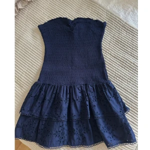 Marinblå klänning - Supersöt marinblå axelbandslös klänning med smockad överdel och volangkjol. Kjolen har broderade blommor och spetsdetaljer längst ner. Väldigt fint skick! Storlek L men passar även M.