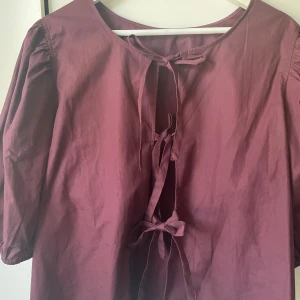 Vinröd blus med knyt och puffärm - Vinröd blus med tre knytband framtill och puffiga trekvartsärmar. Blusen har en lös passform och är tillverkad i bomull, vilket gör den både luftig och bekväm. Perfekt för dig som gillar en romantisk och trendig stil.