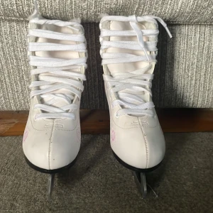 Vita konståkningsskridskor Hudora 35 - Snygga vita konståkningsskridskor från Hudora med ljusrosa blommönster på sidorna. Skorna har snörning, fluffigt foder och klassisk form med svart klack. Perfekta för vinterns isbanor och ger en stilren look på isen. Inte använd mer än 5 gånger. Den är i bra skick men kommer ej med box.