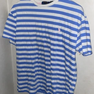 t-shirt Polo Ralph Lauren - Blåvit randig t-shirt från Polo Ralph Lauren med klassisk passform. T-shirten har korta ärmar, rund halsringning och en liten broderad logga på bröstet. Tillverkad i mjuk bomull, perfekt för en chill och stilren look. Inga hål eller fläckar på tröjan.