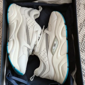 Feta dior skor - Säljer ett par chunky vita Dior sneakers med grå detaljer och blå sula. Skorna har meshpaneler, skinnpartier och reflekterande detaljer som syns i mörker. Snygg och modern siluett med rund tå och snörning. Perfekta för dig som gillar statement sneakers.