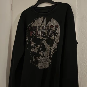 Svart Philipp Plein tröja med döskalle - Cool svart tröja från Philipp Plein i 100% bomull med glittrande döskalle framtill och logga i silver och rött. Långärmad modell med ribbade muddar och relaxed fit. Perfekt statement piece för dig som gillar edgy stil. Pris kan diskuteras ❤️❤️