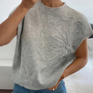 Grå stickad ärmlös tröja - Superfin grå stickad tröja med ärmlös design och stor broderad blomma framtill. Tröjan har ribbad halsringning och nederkant. Använd några gånger men inget som syns fortfarande som ny och lika mjuk.