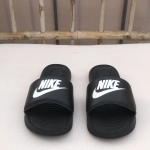 Svarta Nike Tofflor - Svarta tofflor från Nike med stor vit logga över foten. Knappt används. Snygg och enkel design med räfflad sula för extra grepp. Perfekta att glida runt i på sommaren eller till och från träningen.