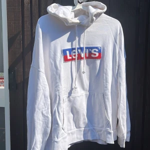 Vit hoodie från Levi's med logga - Vit hoodie från Levi's med stor broderad logga i blått och rött på bröstet. Klassisk känguruficka, snörning i huvan och relaxed fit. Perfekt för dig som gillar streetstyle och vill ha en clean look med tydlig branding.