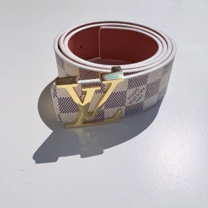 Louis Vuitton rutigt bälte med LV-spänne - Snyggt bälte från Louis Vuitton med klassiskt rutigt mönster i vitt och grått. Bältet har ett stort guldfärgat LV-spänne och insidan är i en brun nyans. Perfekt accessoar för att lyfta din outfit med en lyxig touch.