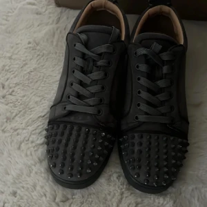 Svarta Louboutin sneakers med nitar - Säljer ett par svarta sneakers från Christian Louboutin i mocka med coola svarta nitar över tån. Skorna har rund tå, platt sula och snörning. Insidan är beige och de har en lyxig vibe som sticker ut. Perfekta för dig som gillar statement-detaljer.